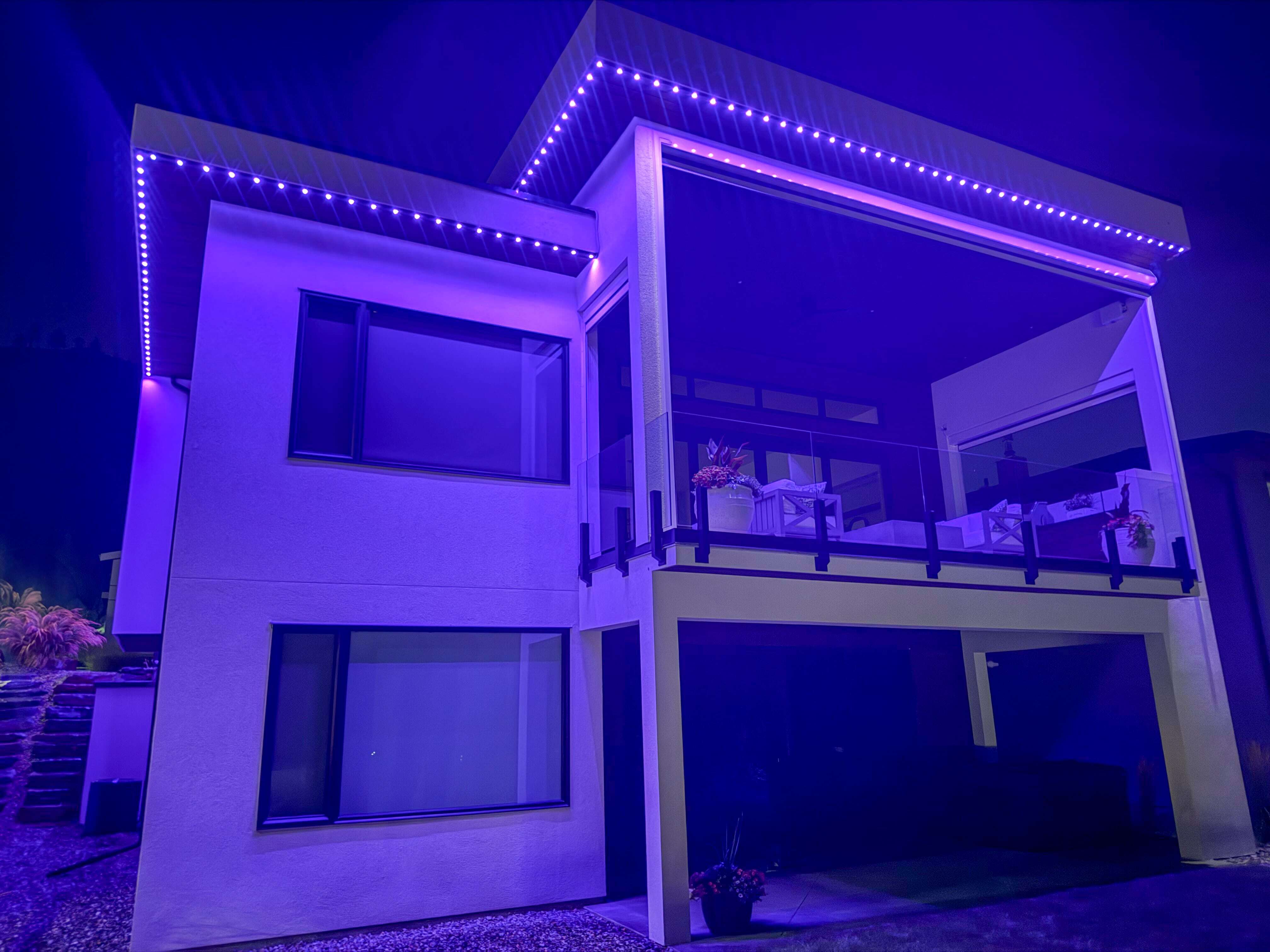 Back view of a modern two-story home illuminated with permanent magenta LED track lighting. The lights outline the roofline and patio, casting a vibrant glow across the walls, windows, and outdoor seating area