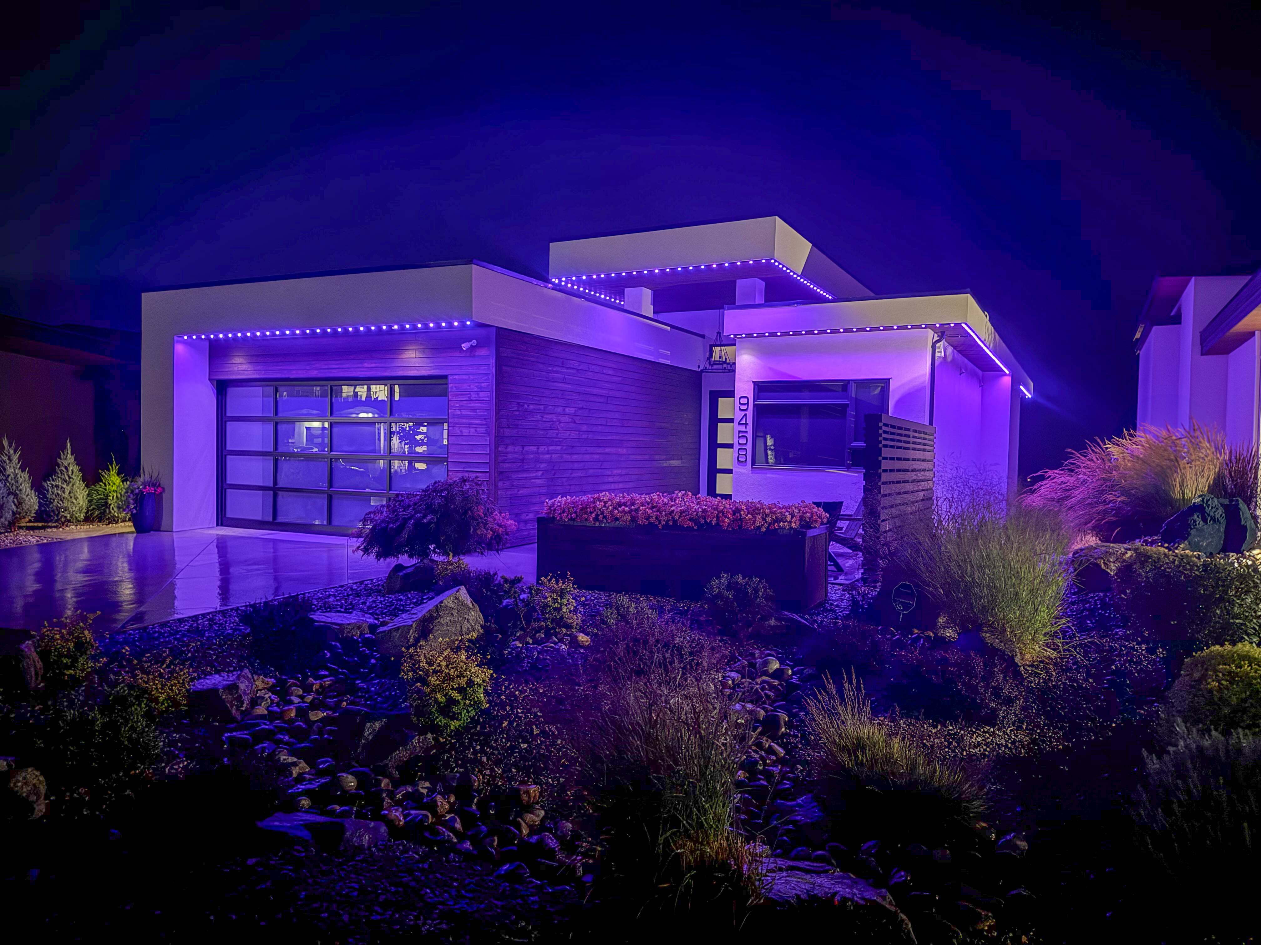 A contemporary home lit with vibrant magenta-purple Perma-Lights outlining the roofline and garage, creating a striking nighttime ambiance with landscaped rocks and plants in the foreground.